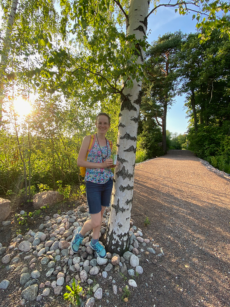 A woman in a flower top and jeans shorts standing by a birch tree.