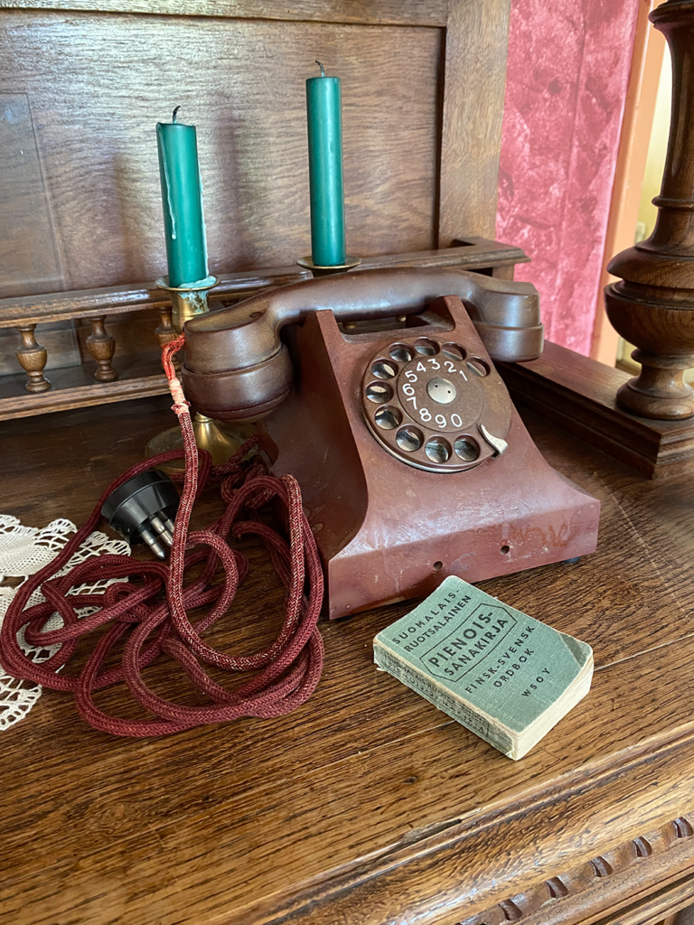 And old brown phone with a long red cord. There is a light green miniature dictionary in front of the phone, and two green candles in candle sticks behind the phone.